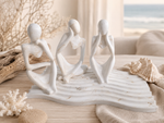 Three abstract seated female figurines in a monochrome off-white finish, arranged on a wave-like base with beach-themed decor including driftwood, coral, and a seashell in a softly lit indoor setting.