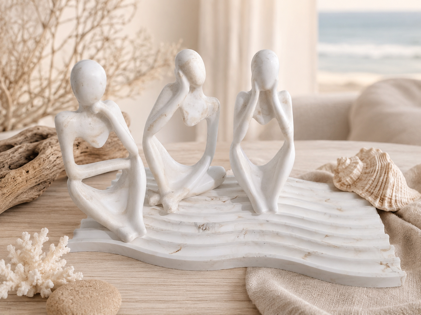 Three abstract seated female figurines in a monochrome off-white finish, arranged on a wave-like base with beach-themed decor including driftwood, coral, and a seashell in a softly lit indoor setting.
