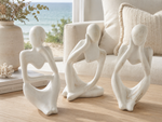 Three minimalist white ceramic figurines of seated women with heart-shaped torsos, displayed on a light wood table in a bright living room with a beach view in the background.