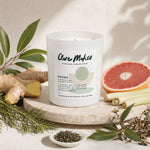 A white glass jar candle labeled 'Clare Makes' is styled on a stone slab with botanical ingredients including ginger, lemongrass, green leaves, and a grapefruit slice on a light neutral background.
