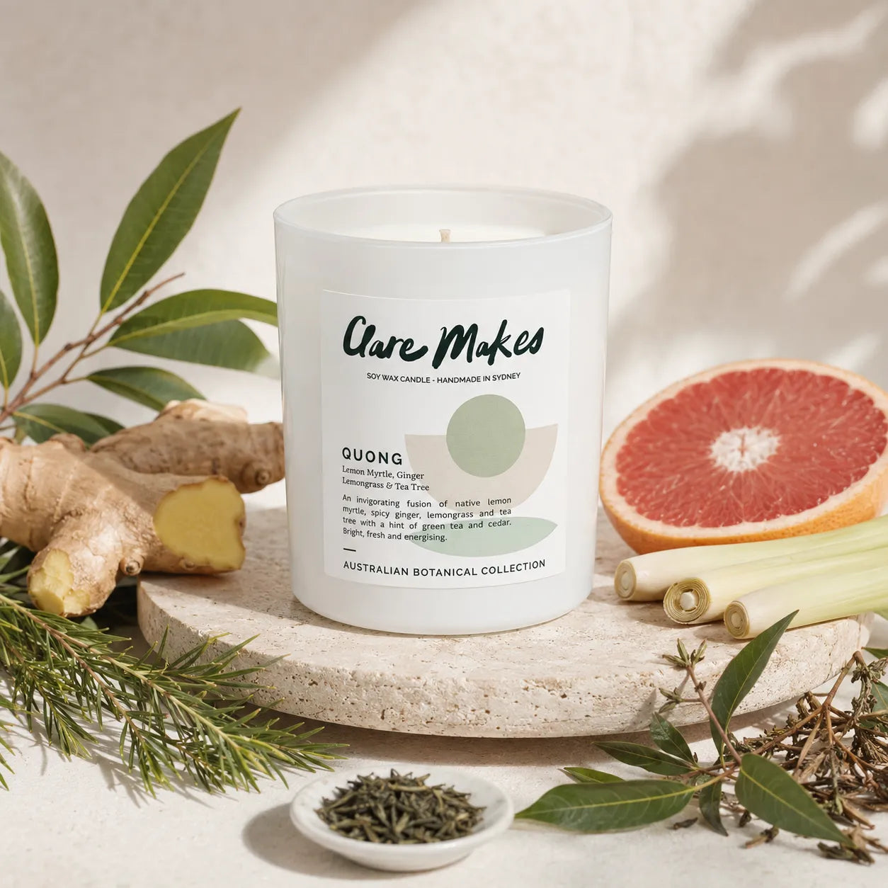 A white glass jar candle labeled 'Clare Makes' is styled on a stone slab with botanical ingredients including ginger, lemongrass, green leaves, and a grapefruit slice on a light neutral background.