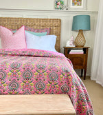 Bedroom with pink patterned bedding, wooden headboard, and nightstand with lamp.