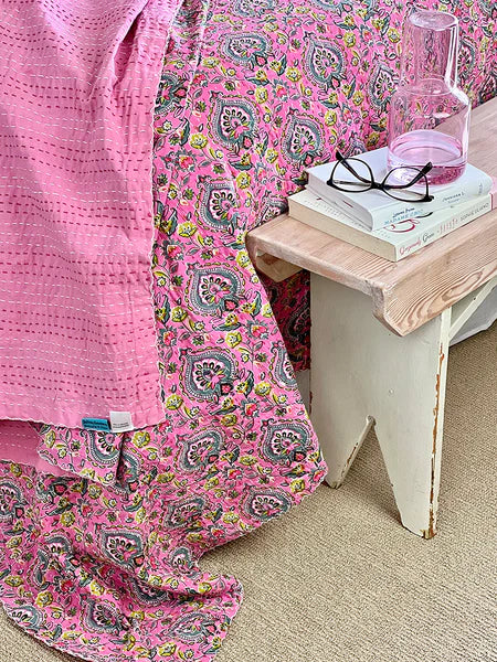 Pink floral-patterned blanket draped over a bed with a small wooden table holding books and glasses.