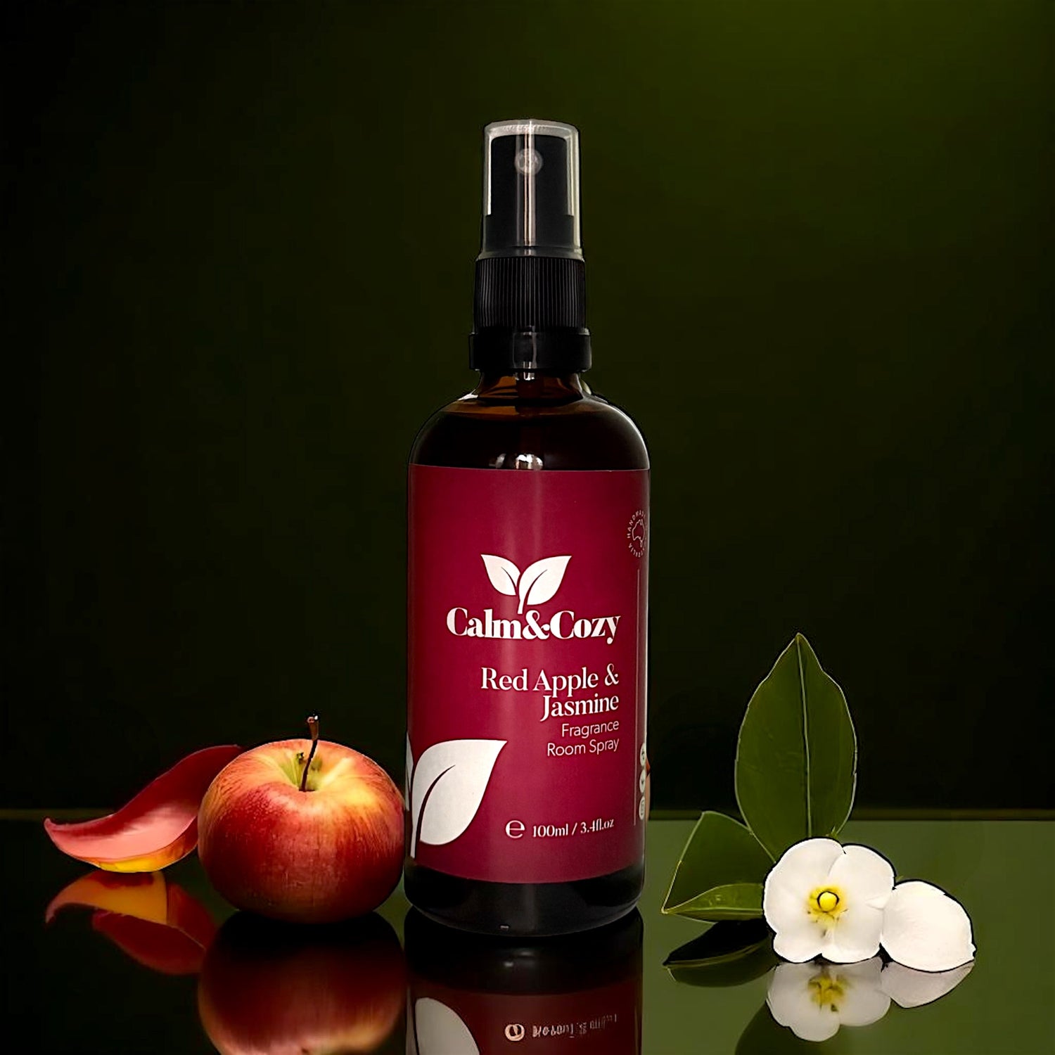 Bottle of Calm & Cozy Red Apple & Jasmine fragrance room spray with an apple and flowers on a reflective surface.