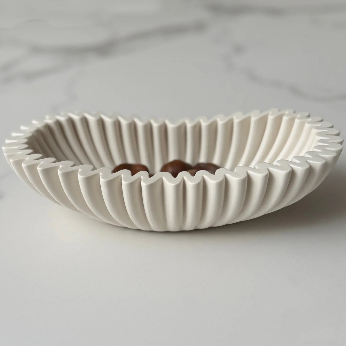 A white ceramic bowl with a fluted, scalloped rim, shown on a light gray marble surface with three dark brown chocolates inside.