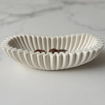 A white ceramic bowl with a fluted, scalloped rim, shown on a light gray marble surface with three dark brown chocolates inside.