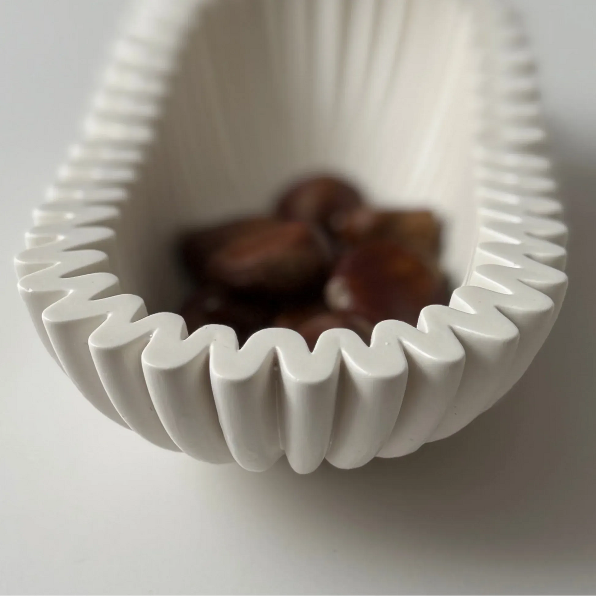 A white ceramic bowl with a wavy, pleated rim, shown close-up on a light gray background with dark brown chestnuts inside.