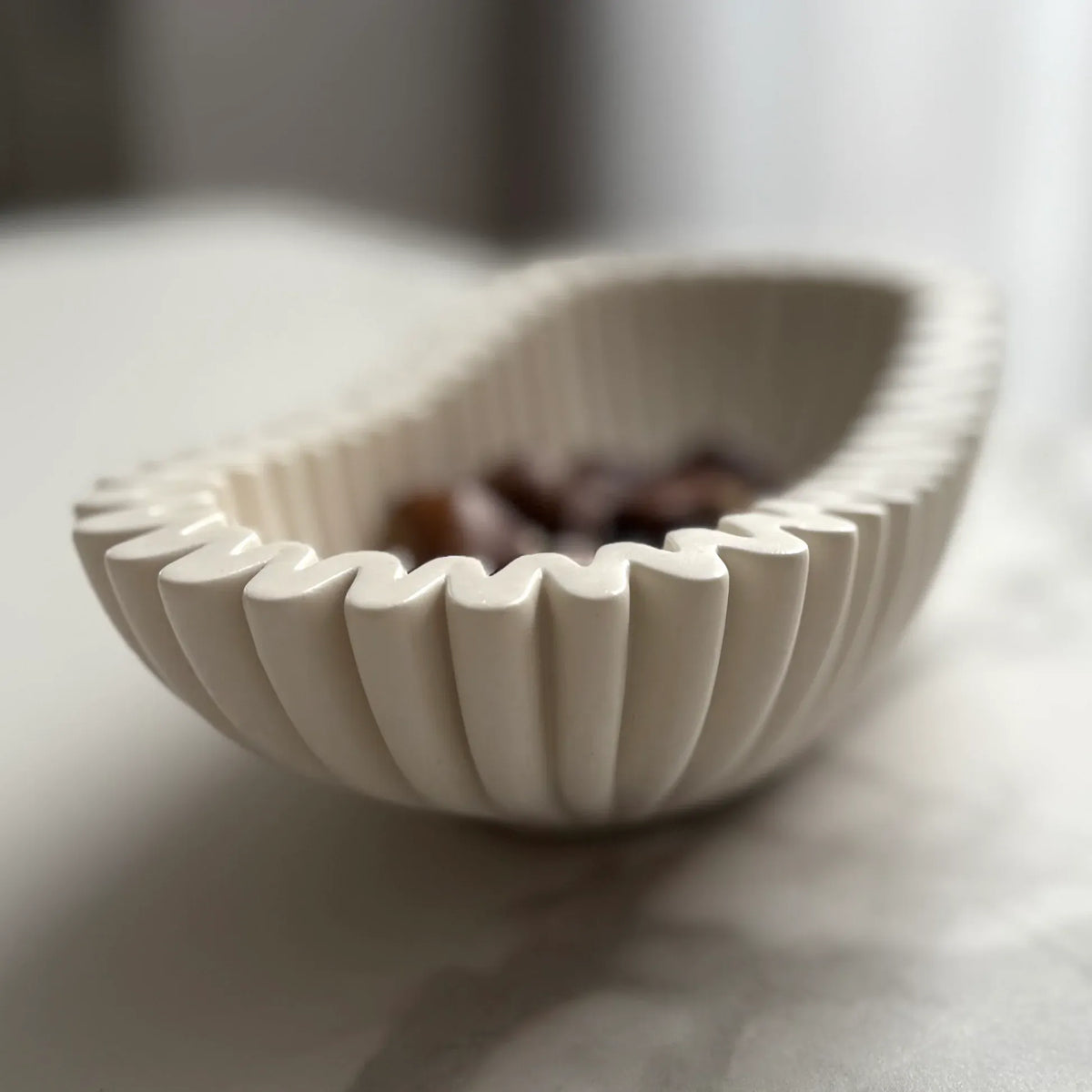 A small off-white ceramic bowl with a fluted, scalloped rim, shown on a light marble-like surface with blurred dark stones inside.