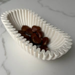 A white ceramic oval dish with a wavy, pleated rim, containing several brown chestnuts, set on a light gray marble surface.