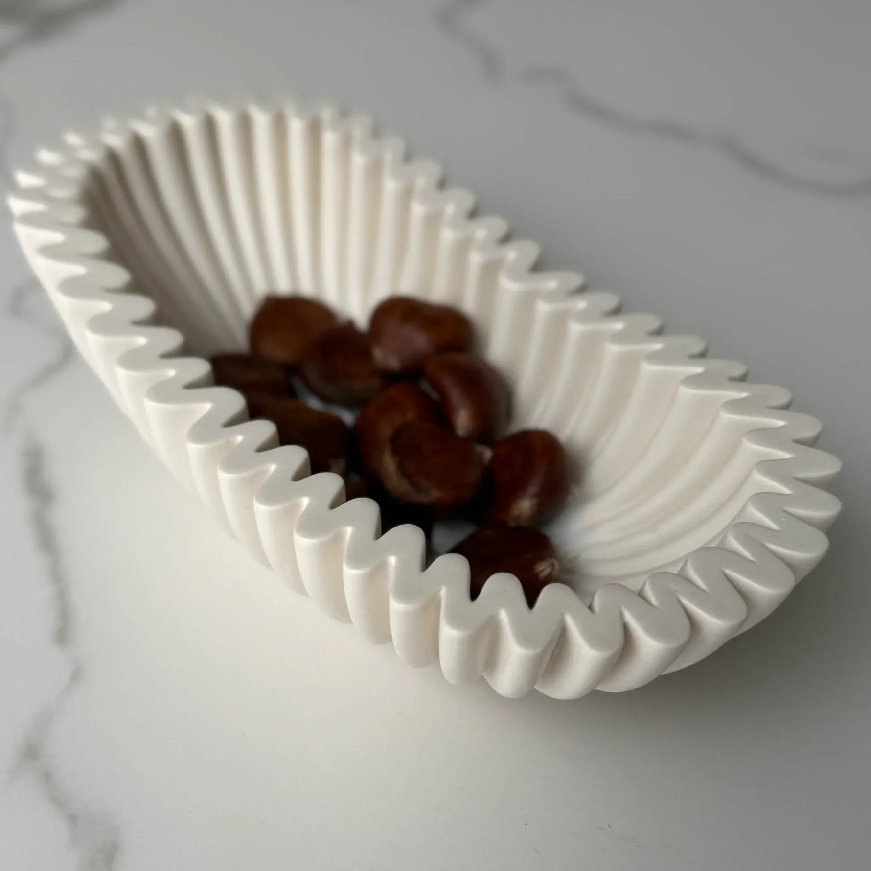 A white ceramic oval dish with a wavy, pleated rim, containing several brown chestnuts, set on a light gray marble surface.