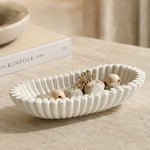 A ribbed oval ceramic bowl in warm off-white, styled with dried botanicals and wooden beads on a beige stone surface, with a blurred book in the background.