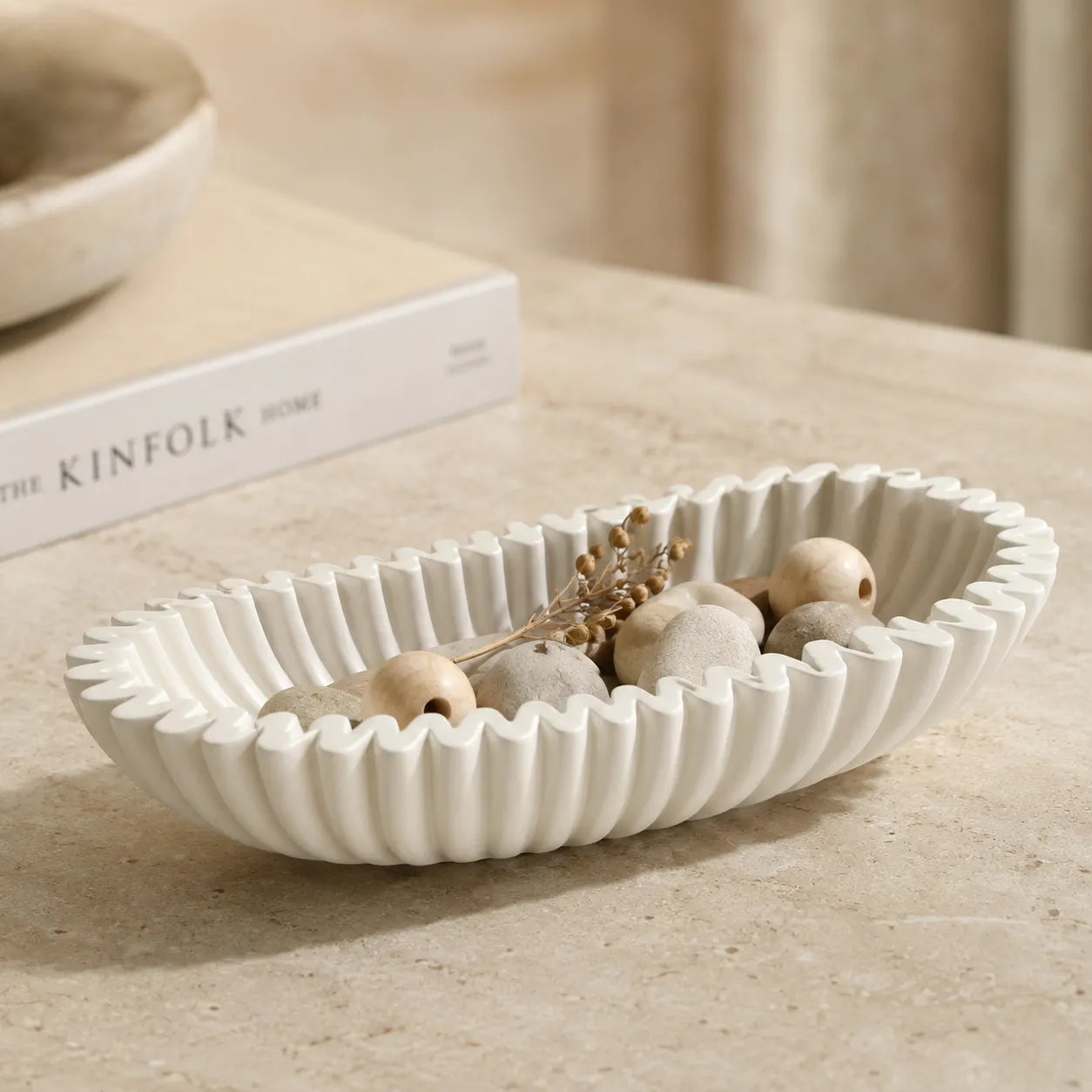 A ribbed oval ceramic bowl in warm off-white, styled with dried botanicals and wooden beads on a beige stone surface, with a blurred book in the background.