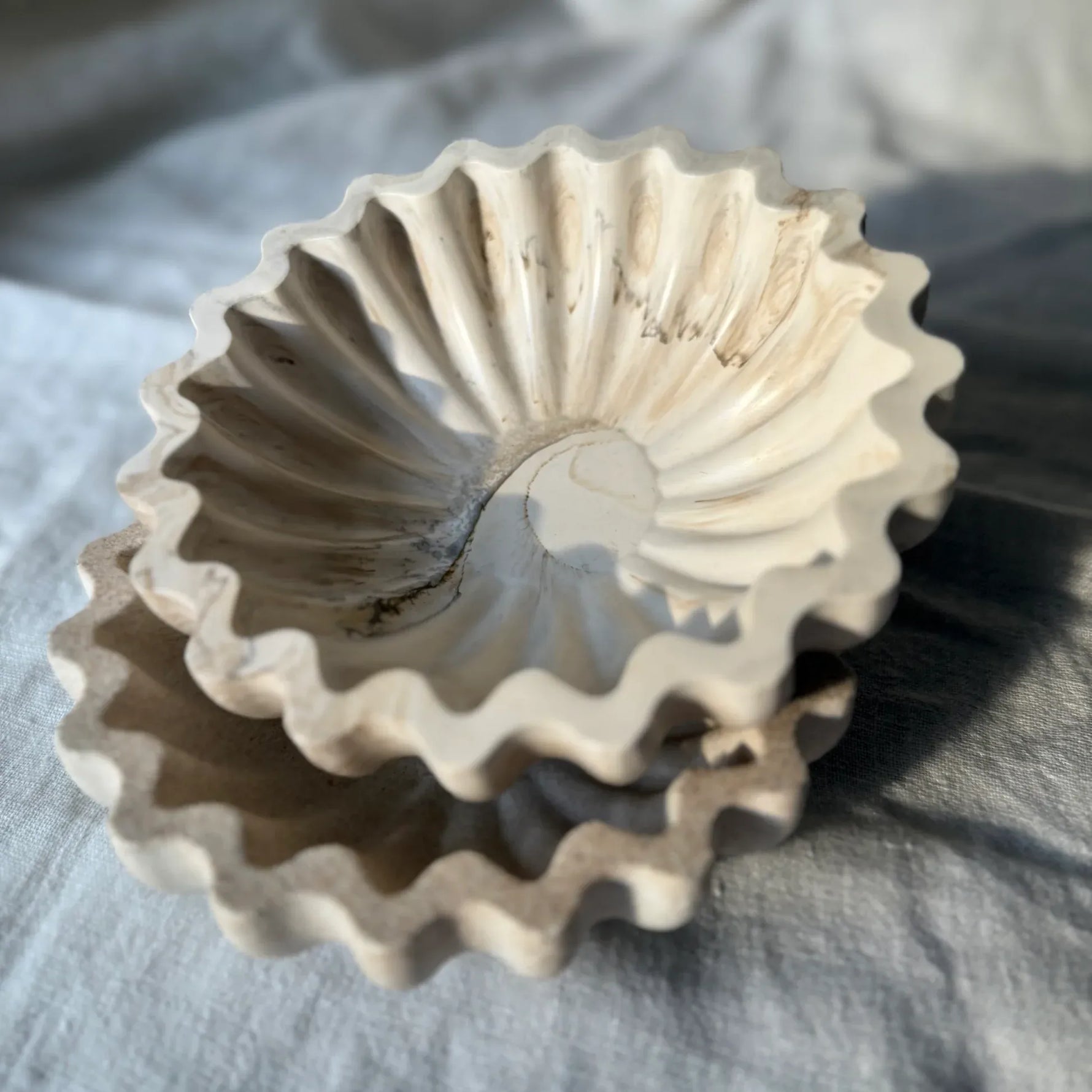 A stack of three scalloped ceramic dishes or bowls in a light warm off-white glaze, resting on a textured gray fabric background.