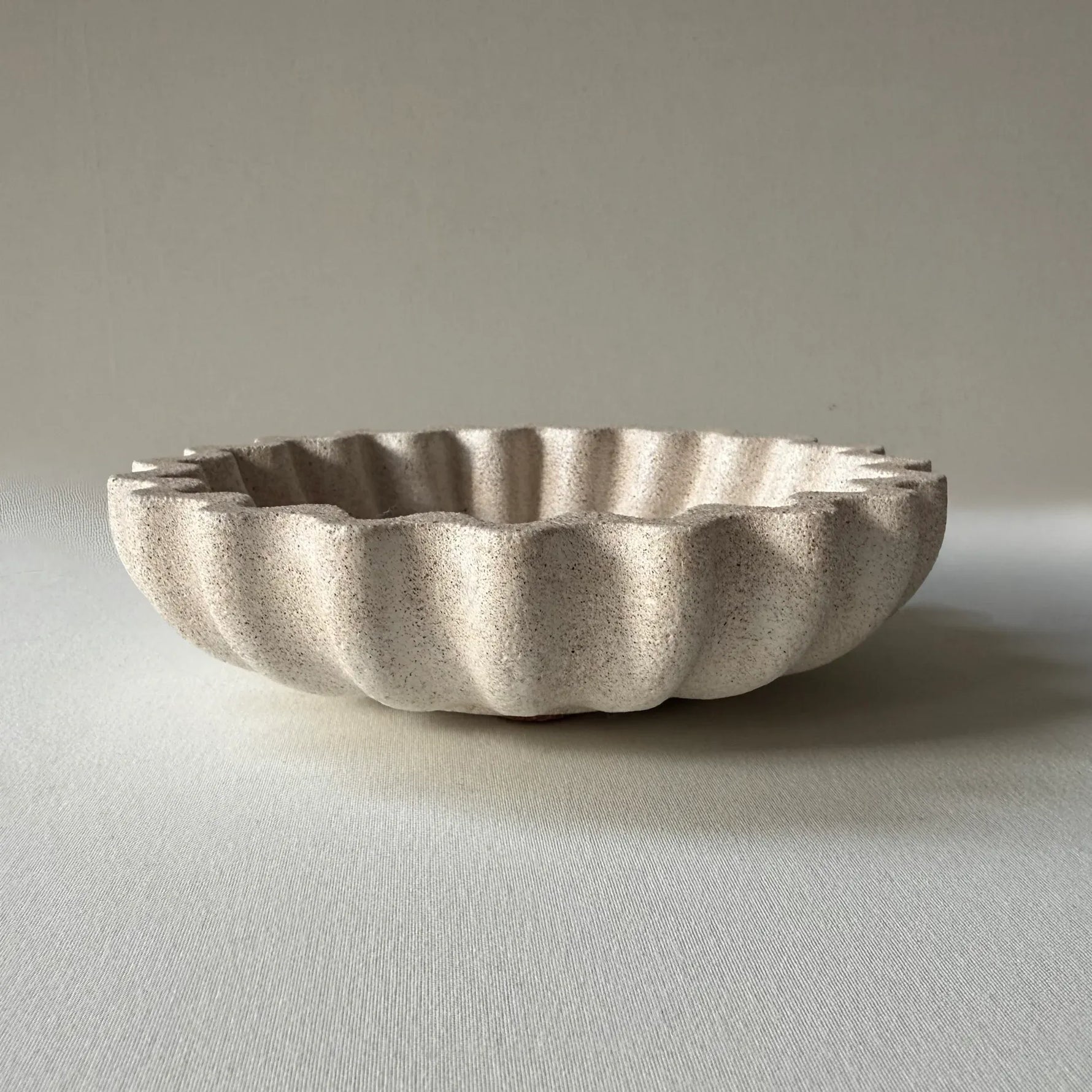 A small scalloped ceramic dish in a speckled light beige or off-white glaze, photographed on a neutral light gray background.