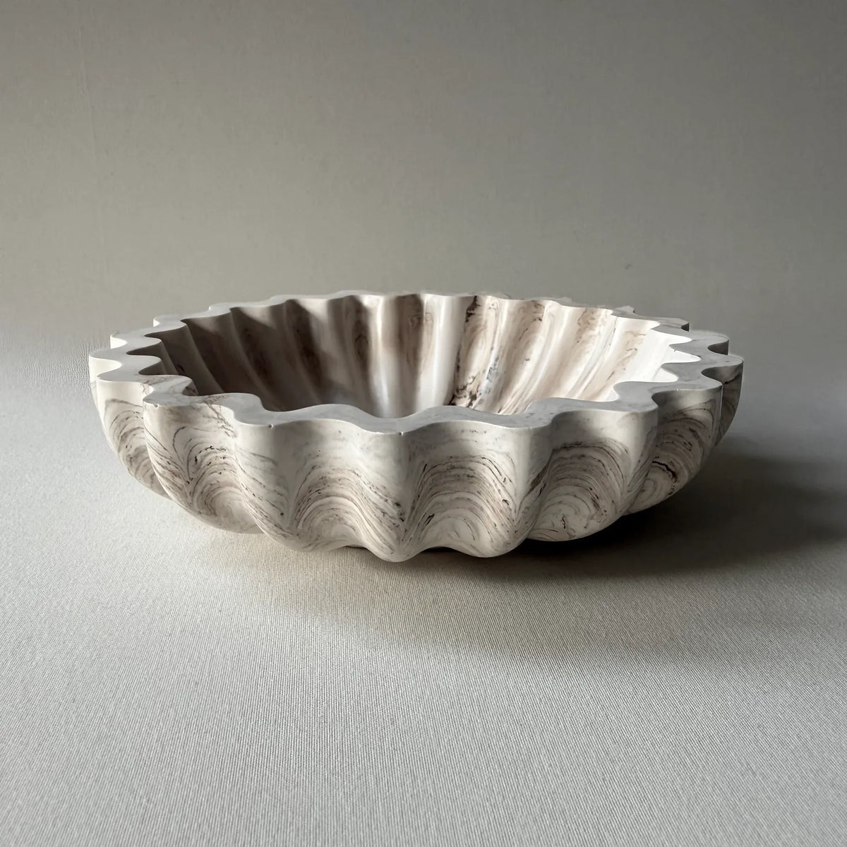 A scalloped ceramic bowl with a fluted rim, featuring a marbled pattern in off-white, beige, and gray, set against a neutral gray background.