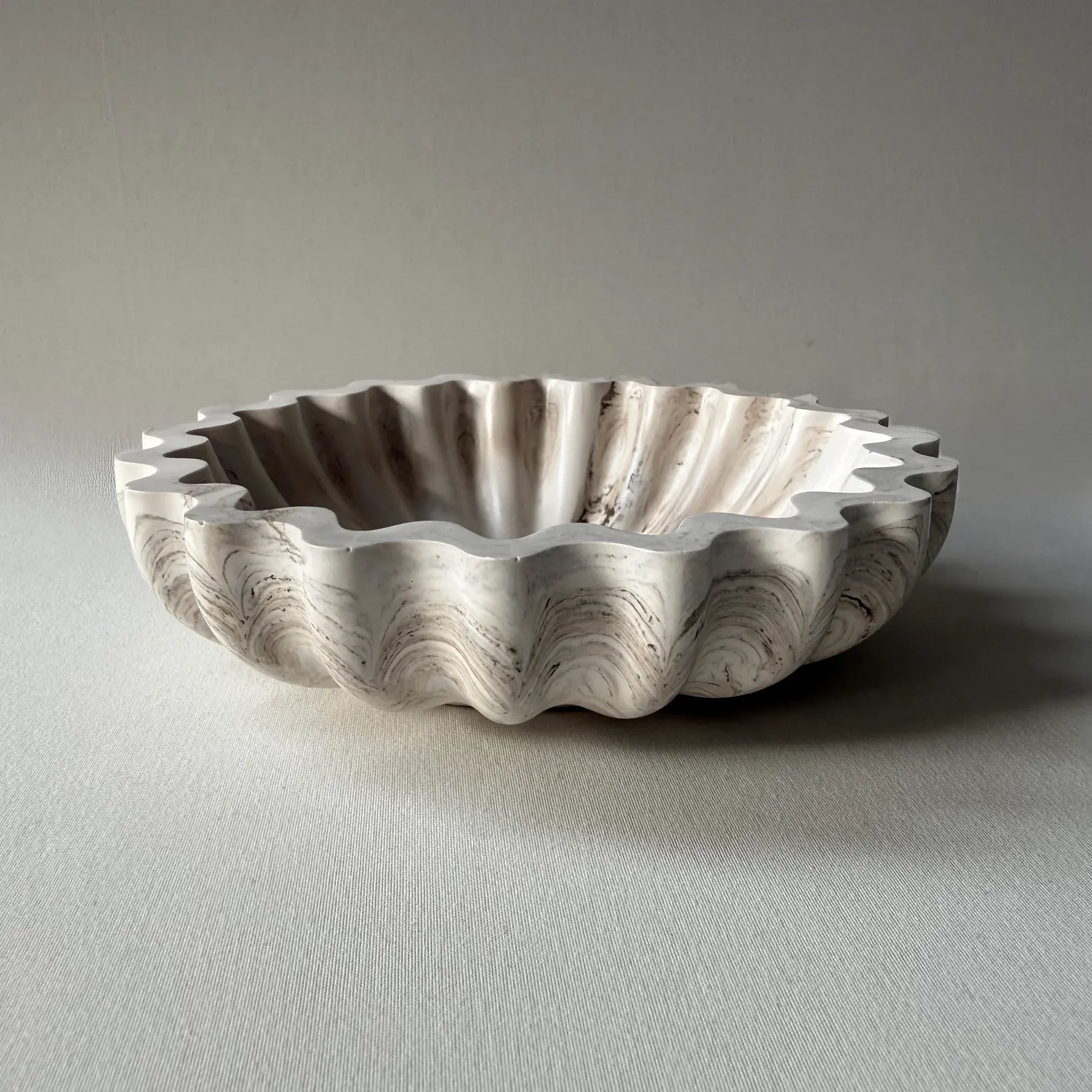 A scalloped ceramic bowl with a fluted rim, featuring a marbled pattern in off-white, beige, and gray, set against a neutral gray background.