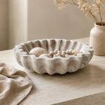 A scalloped ceramic bowl with a textured, stone-like finish in warm off-white and beige, styled on a light stone tabletop with dried flowers and a linen cloth.