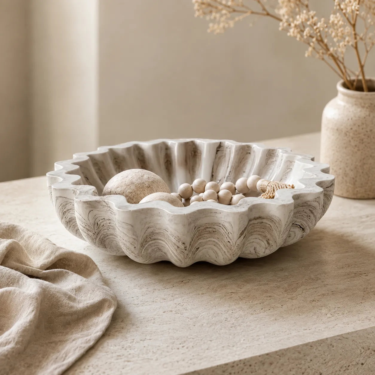 A scalloped ceramic bowl with a textured, stone-like finish in warm off-white and beige, styled on a light stone tabletop with dried flowers and a linen cloth.