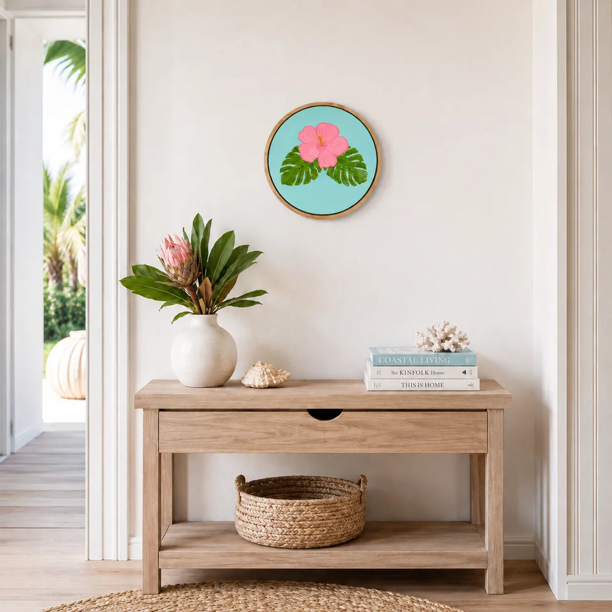 A round framed wall art print featuring a pink hibiscus flower on a light blue background, styled on a beige wall above a light wood console table with a vase of flowers, books, and a woven basket.