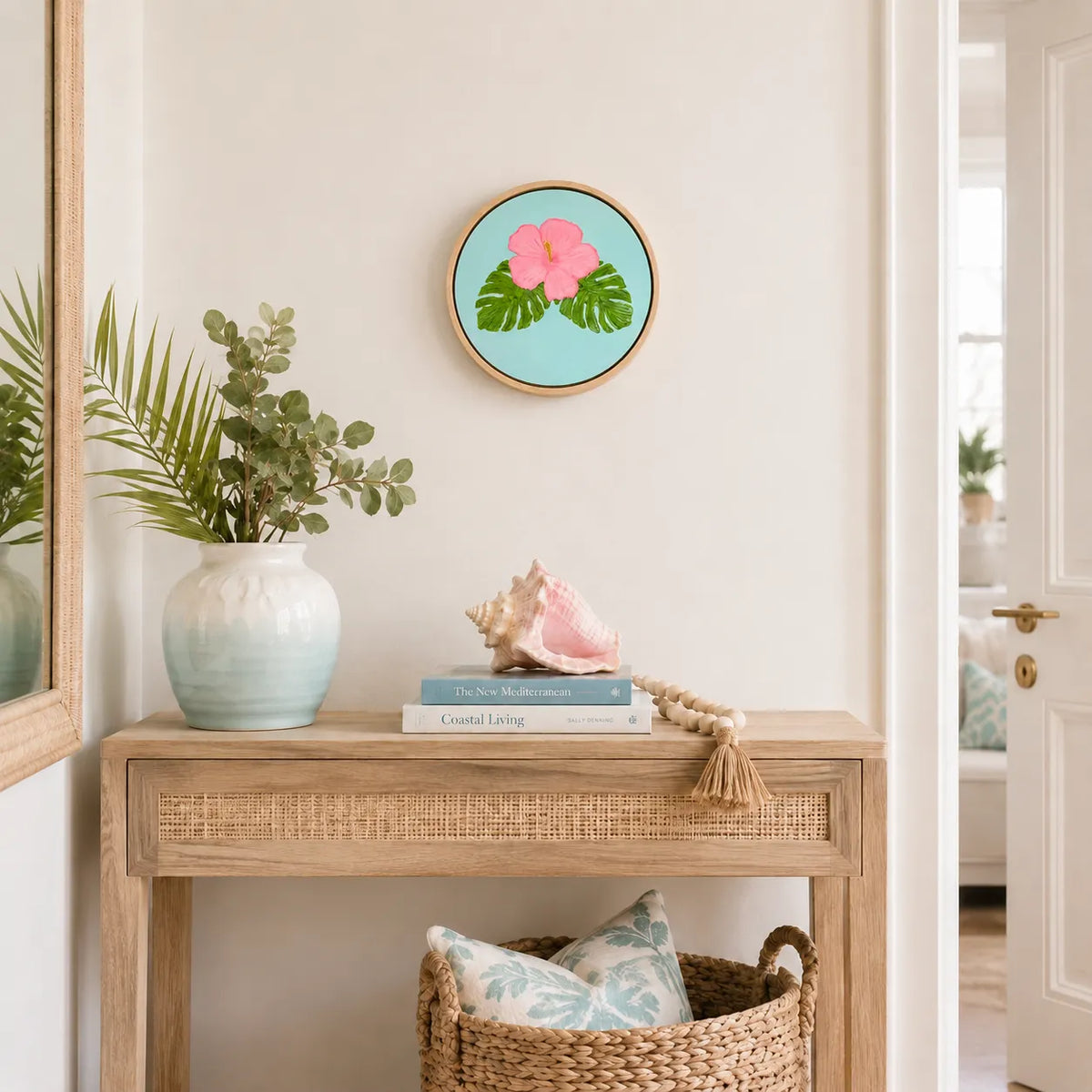 A round framed art print featuring a pink hibiscus flower and green leaves on a light blue background, styled on a light wood console table in a bright, coastal-themed room.