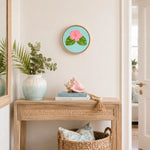 A round framed art print featuring a pink hibiscus flower and green leaves on a light blue background, styled on a light wood console table in a bright, coastal-themed room.