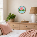 A round embroidered wall art piece featuring a pink hibiscus flower and green monstera leaves on a light blue background, hanging on a beige wall above a wooden dresser in a styled bedroom.
