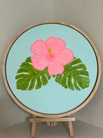 A circular framed painting on an easel, featuring a large pink hibiscus flower and green monstera leaves on a light blue background.
