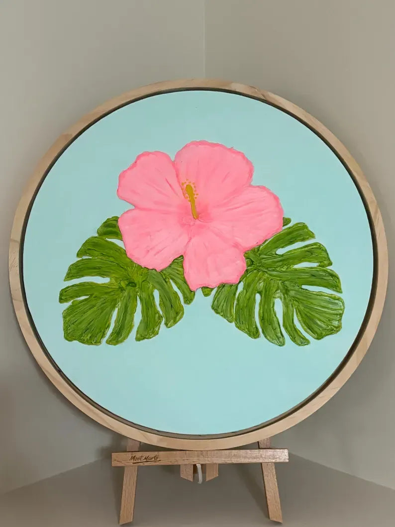 A circular framed painting on an easel, featuring a large pink hibiscus flower and green monstera leaves on a light blue background.