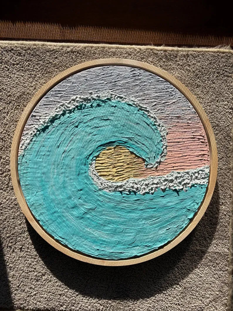 A circular wooden hoop wall art piece featuring a textured, 3D ocean wave design in turquoise, white, pink, and gold, mounted on a textured gray wall.