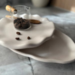 A ceramic coffee table setting featuring a small white wavy plate holding a clear glass pitcher filled with coffee beans, with a few loose beans scattered on the plate, set on a larger matching white plate over a gray marble surface.