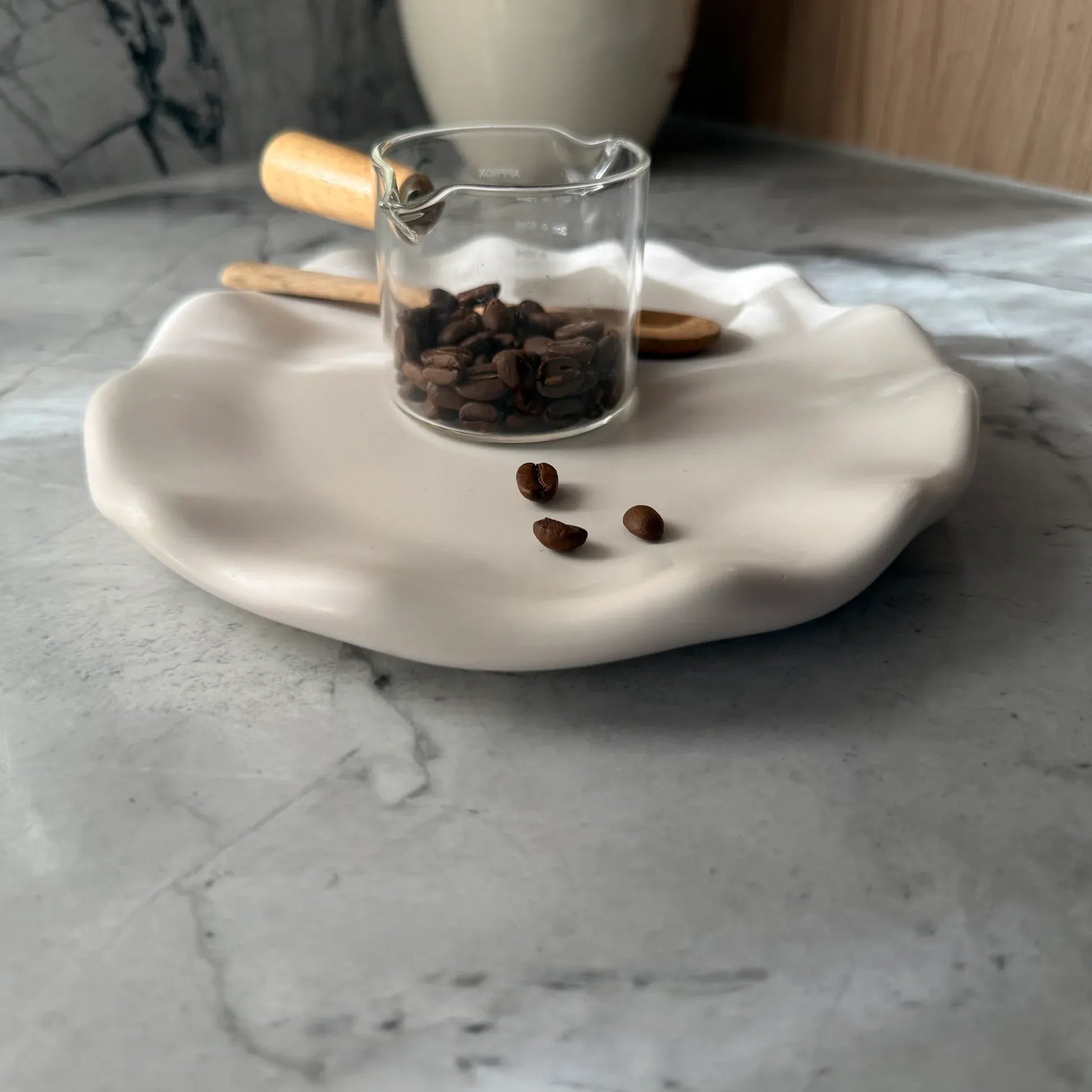 A small clear glass measuring cup filled with dark brown coffee beans sits on a white ceramic plate with a wavy scalloped edge, placed on a gray marble surface.