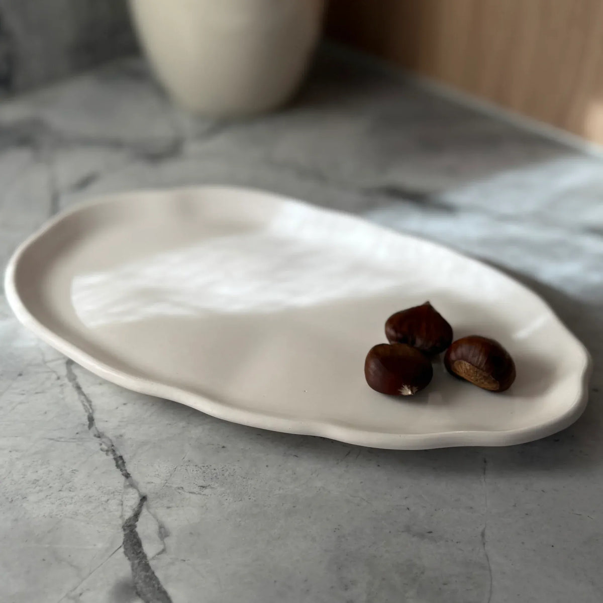 An organic-shaped matte white ceramic plate with scalloped edges, styled on a gray marble surface with three brown chestnuts on top.