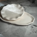 Two off-white ceramic plates with organic, wavy edges, one round scalloped plate resting on top of a larger oval plate, set on a gray marble surface.