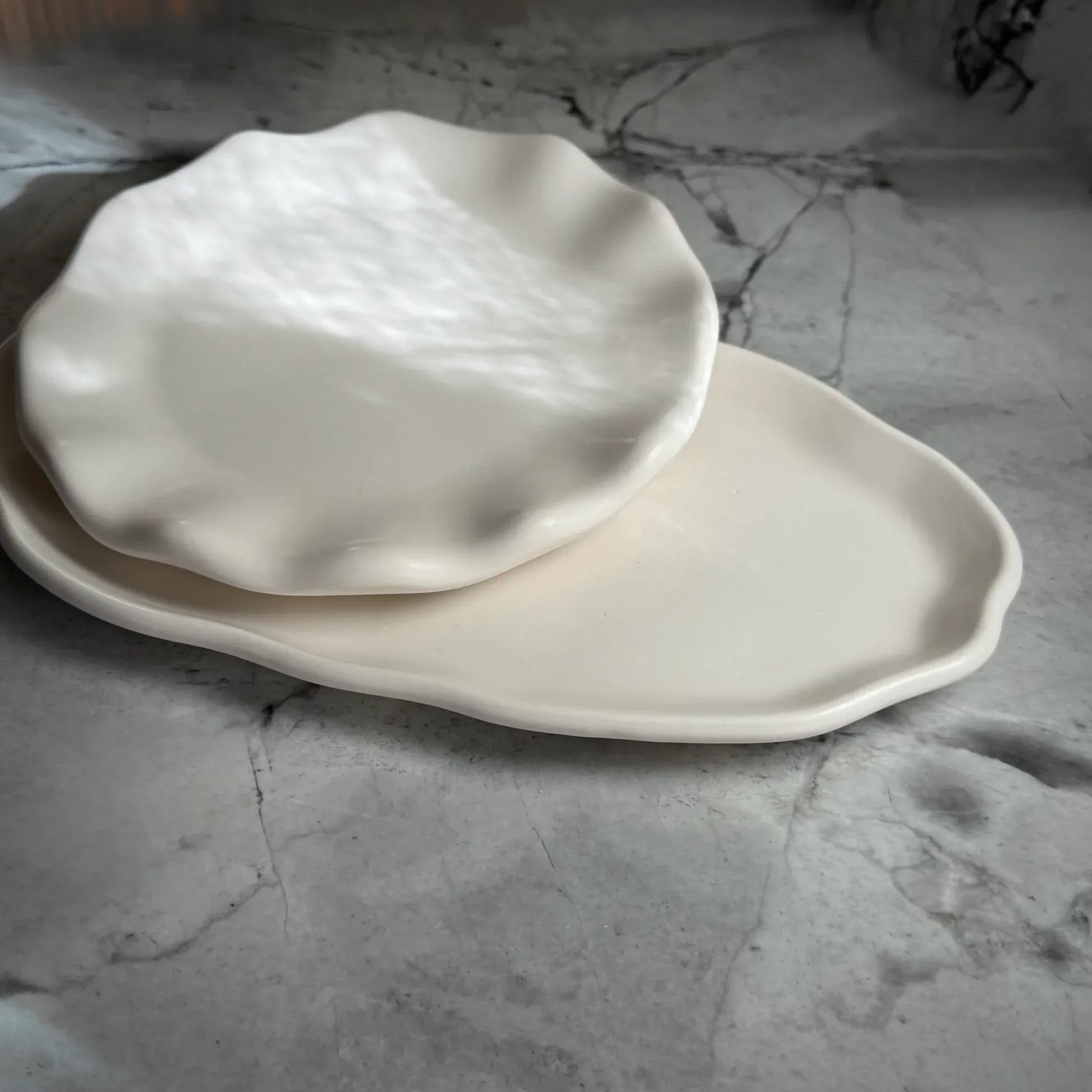 Two off-white ceramic plates with organic, wavy edges, one round scalloped plate resting on top of a larger oval plate, set on a gray marble surface.