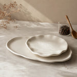 A set of three organic-shaped ceramic plates in a warm off-white color, styled on a light marble tabletop with dried flowers, a small glass jar of coffee beans, and a wooden spoon.