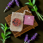 Beige soap in a mesh bag with a purple box labeled 'Calm & Cozy' on a dark surface with lavender flowers.