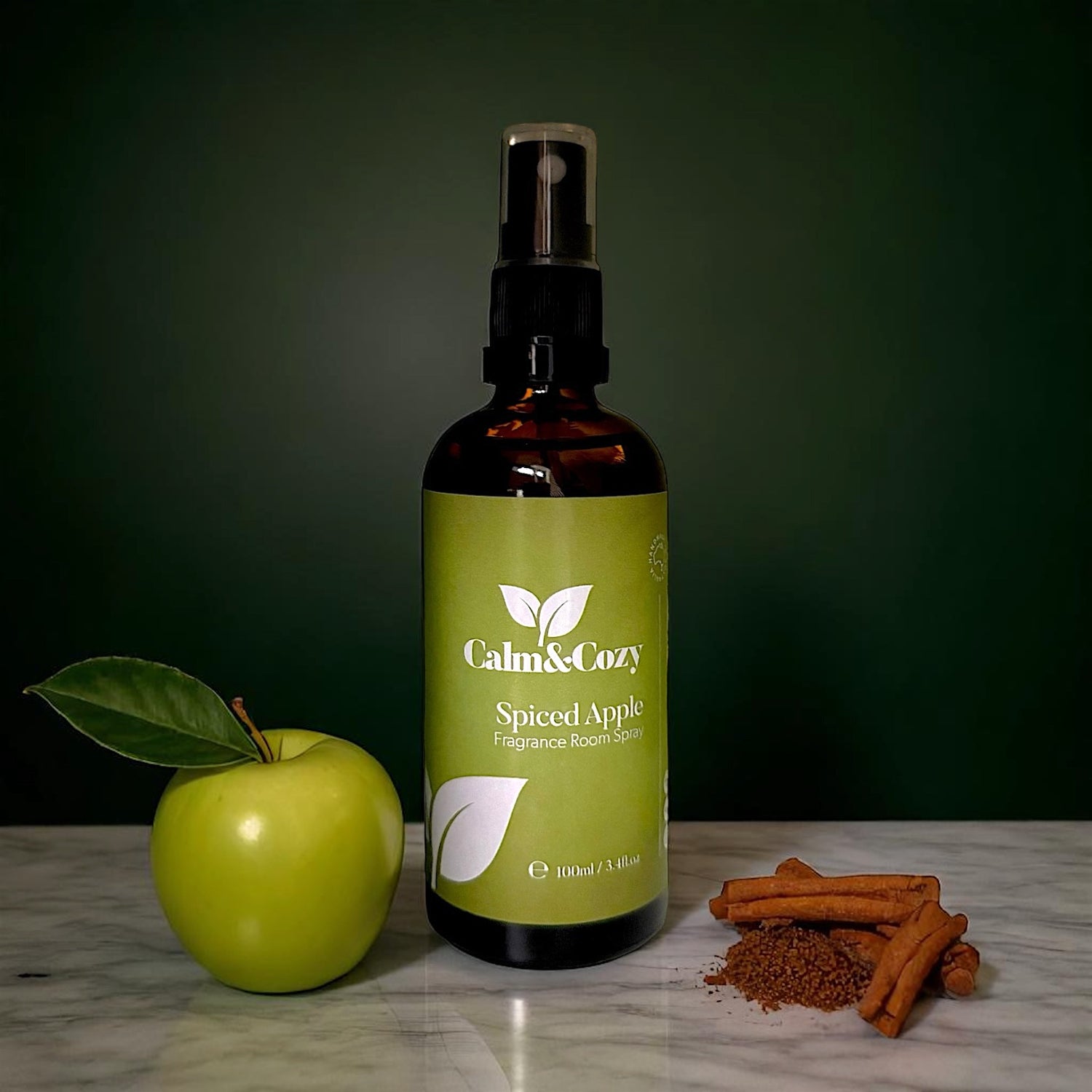 Bottle of Calm & Cozy Spiced Apple fragrance room spray with an apple and cinnamon sticks on a marble surface.