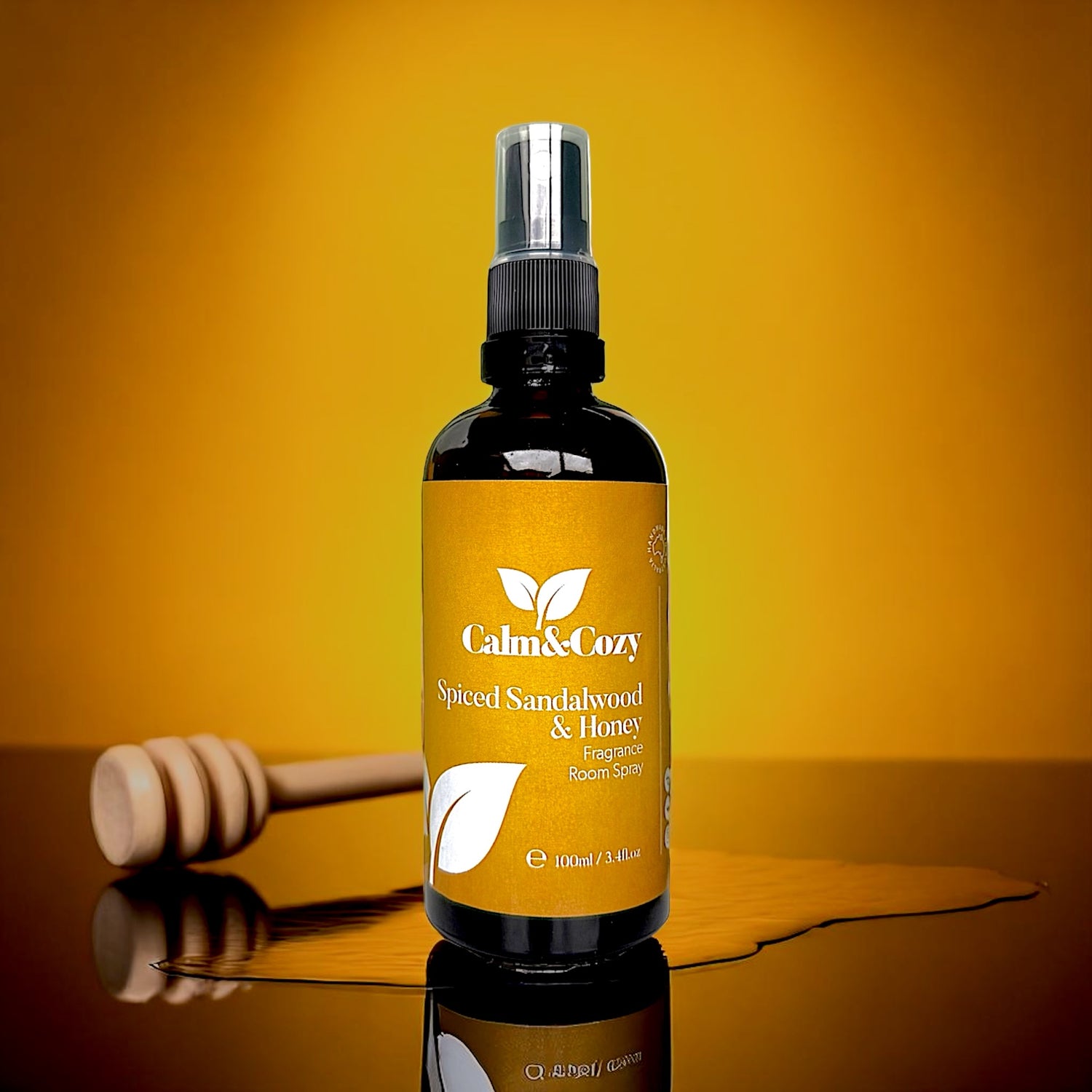 Bottle of 'Calm & Cozy' Spiced Sandalwood & Honey product on a reflective surface with a wooden honey dipper.