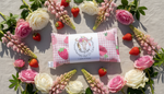 Pink and white checkered  heat pack with strawberry design surrounded by flowers and strawberries on a wooden surface.