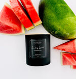 A black scented candle jar labeled 'Luna Bleu Collection Sunday Lunch Watermelon' is styled on a white surface with watermelon slices and a whole watermelon around it.