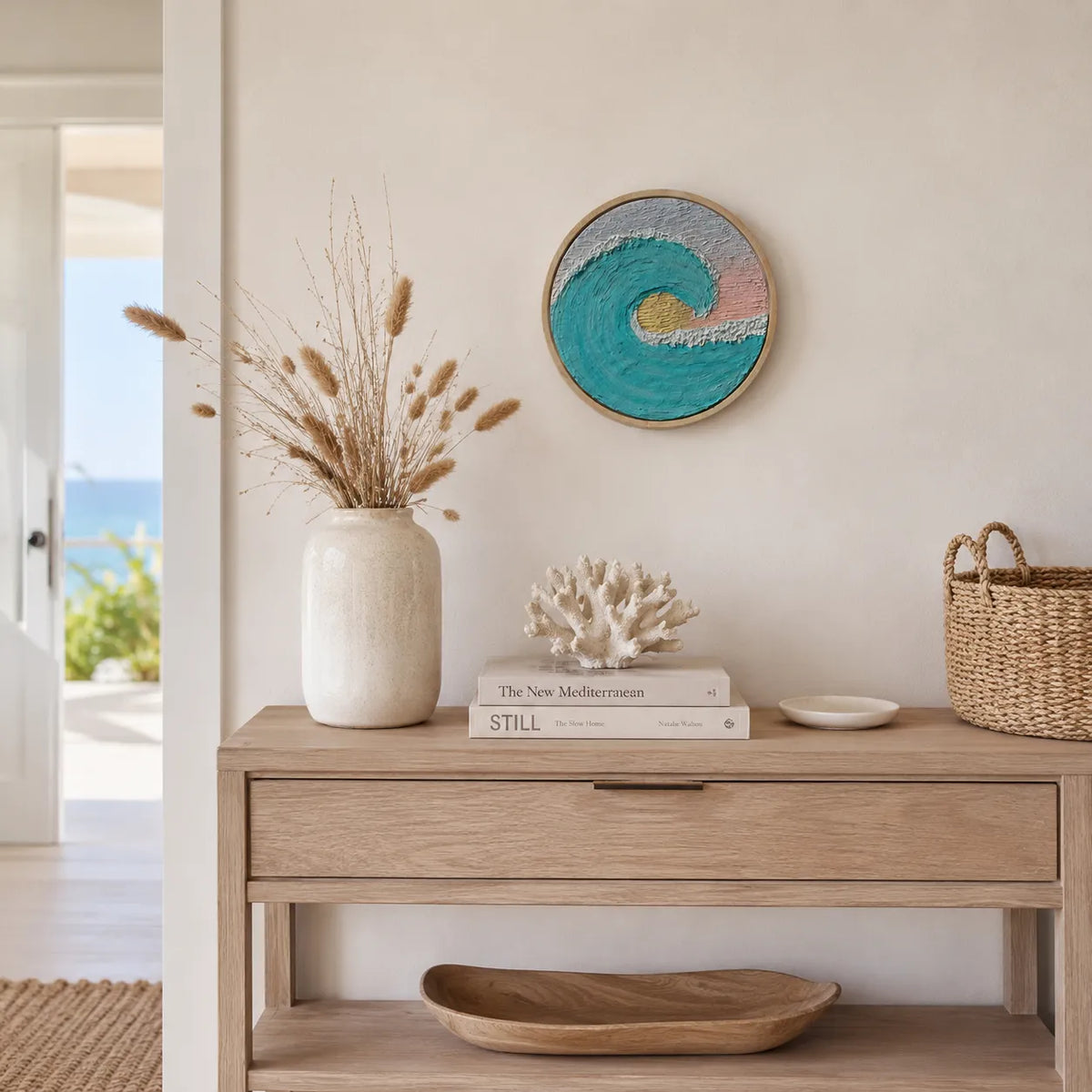 A round wooden-framed wall art piece with a turquoise, white, and pink ocean wave design hangs on a beige wall above a light wood console table styled with a vase of dried grasses, books, a coral sculpture, and a woven basket.