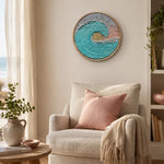 A round framed wall art piece featuring a textured turquoise, white, and soft pink ocean wave hangs above a beige armchair in a softly styled room with a pink pillow and throw blanket.