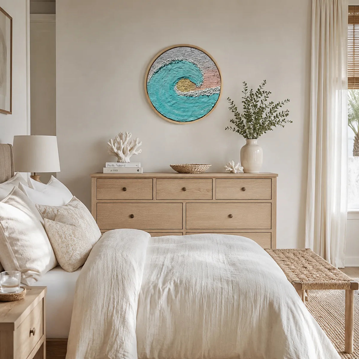 A styled bedroom featuring a light wood dresser with decorative items, a bed with white linens, and a round framed wall art piece depicting a turquoise ocean wave above the dresser.