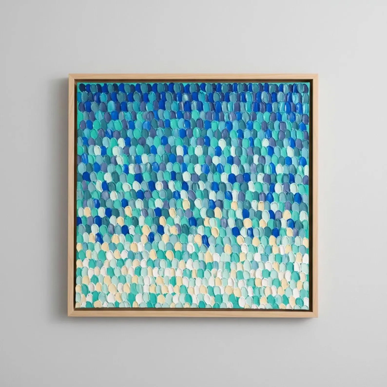 A square framed abstract wall art piece featuring a textured pattern of overlapping pebble-like shapes in shades of blue, teal, aqua, and cream, mounted on a light gray wall.