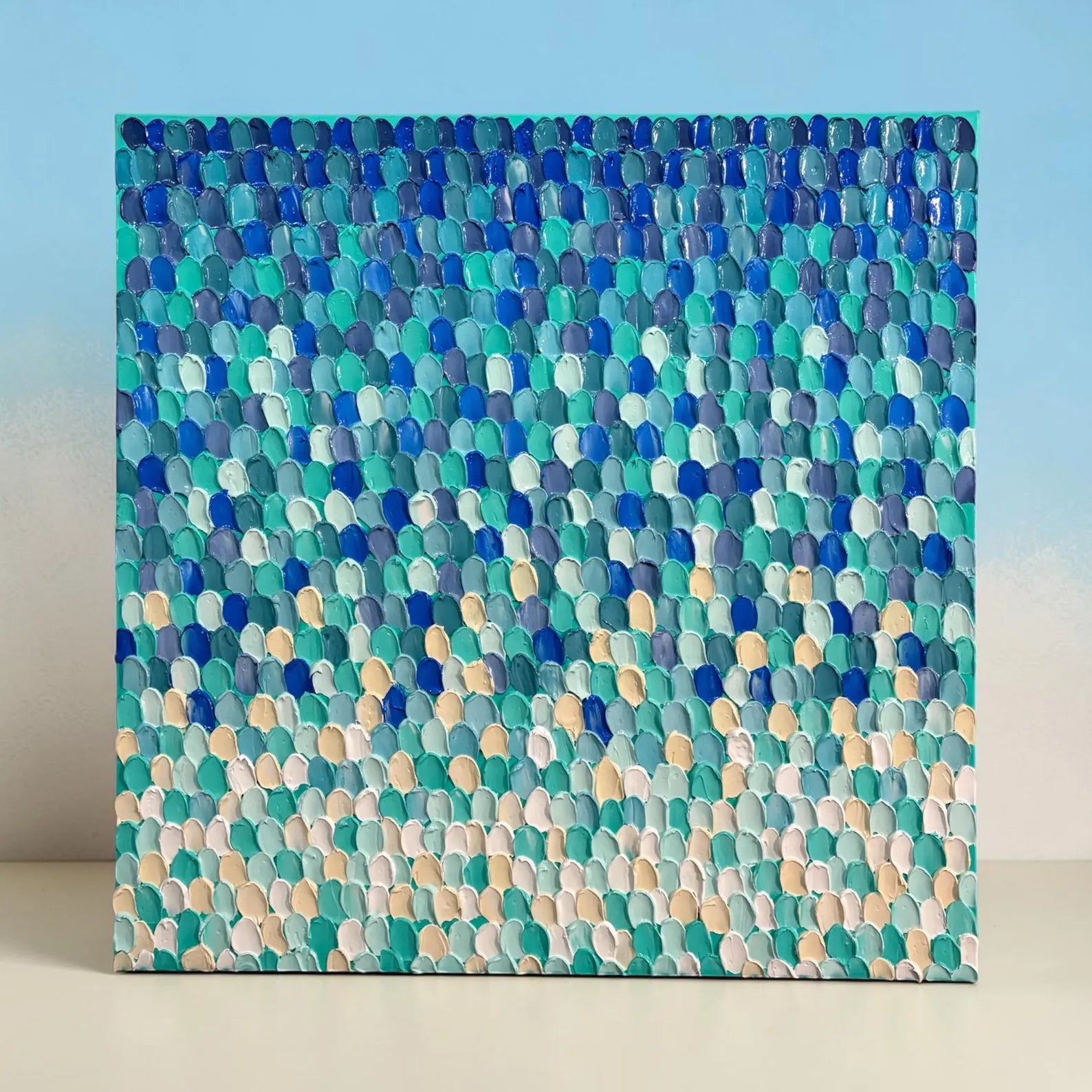 A square canvas painting featuring a textured, impasto pattern of overlapping pebble-like shapes in shades of blue, teal, green, and beige, displayed against a light blue and white background.
