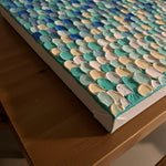 A close-up of a textured canvas wall art piece with thick, raised paint strokes in shades of teal, blue, mint green, cream, and white, resting on a wooden table.