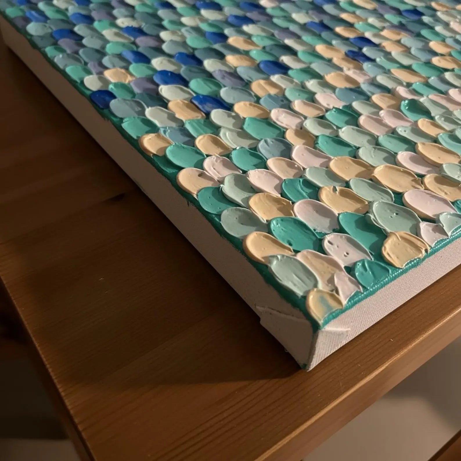 A close-up of a textured canvas wall art piece with thick, raised paint strokes in shades of teal, blue, mint green, cream, and white, resting on a wooden table.