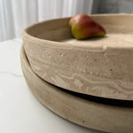 Two stacked round ceramic plates in a matte, textured beige finish with a small red and green fruit resting on the top plate, set on a white marble surface.