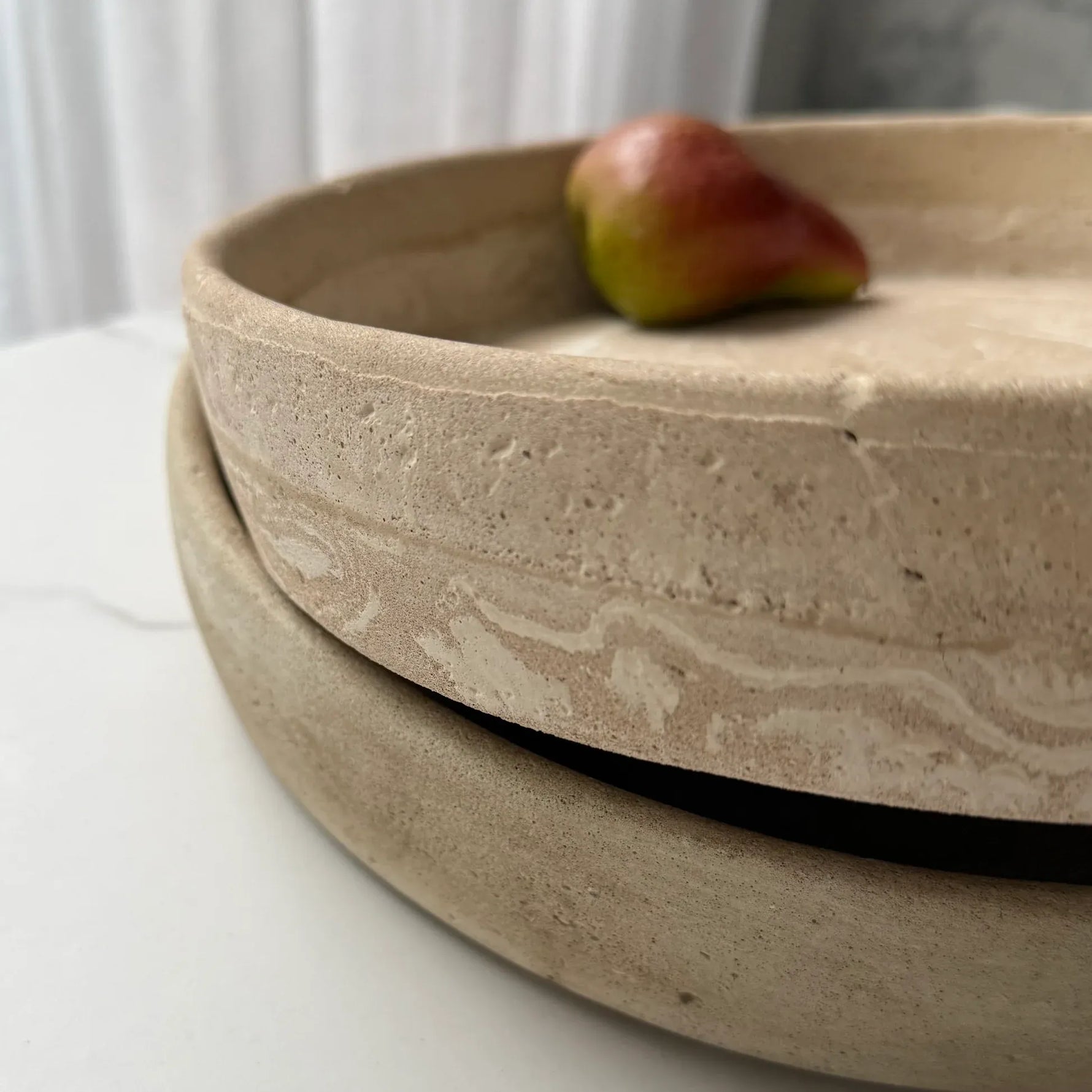 Two stacked round ceramic plates in a matte, textured beige finish with a small red and green fruit resting on the top plate, set on a white marble surface.