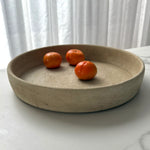 A round, shallow concrete tray with a speckled beige finish, styled with three small orange fruits on a light marble surface in front of white curtains.